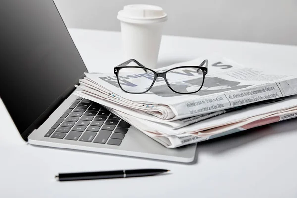Messa a fuoco selettiva del computer portatile con schermo bianco vicino a giornali aziendali, occhiali, penna e tazza di carta su bianco — Foto stock