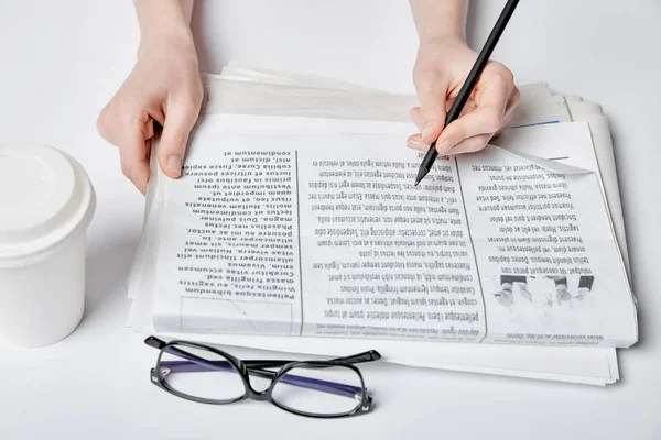 Cropped view of woman holding pencil near newspaper, glasses and paper cup with drink on white — Stock Photo