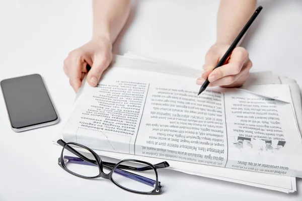 Vista recortada de la mujer sosteniendo lápiz cerca de periódico, gafas y teléfono inteligente con pantalla en blanco - foto de stock