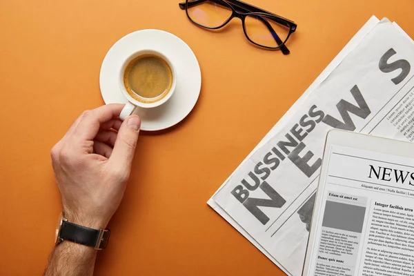 Abgeschnittene Ansicht eines Mannes, der Tasse mit Kaffee in der Nähe von Gläsern und Wirtschaftszeitung auf Orange hält — Stockfoto