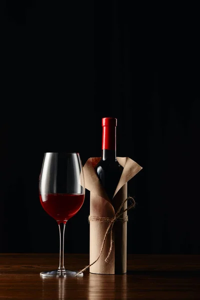 Wine glass and bottle in paper wrapper on wooden surface isolated on black — Stock Photo