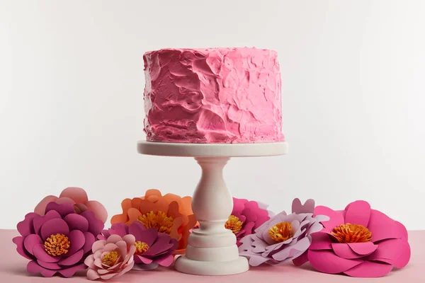 Gâteau d'anniversaire rose sur le stand de gâteau près de fleurs en papier isolé sur gris — Photo de stock