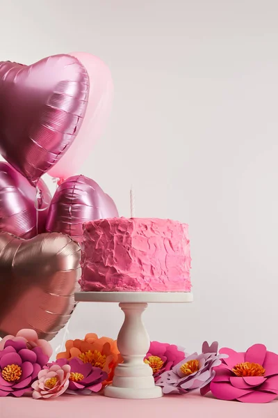 Pink birthday cake with candle on cake stand near paper flowers and heart-shaped air balloons on grey — Stock Photo
