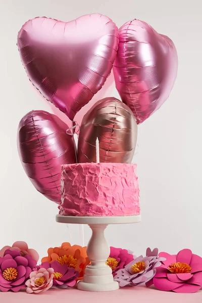 Gâteau d'anniversaire rose savoureux avec bougie sur le stand de gâteau près des fleurs en papier et des ballons à air en forme de coeur sur gris — Photo de stock