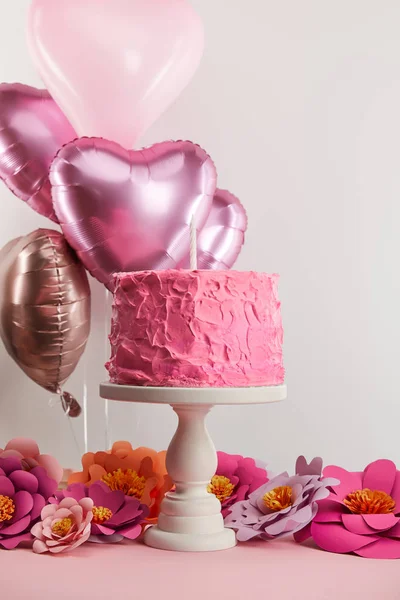 Sweet pink birthday cake with candle on cake stand near paper flowers and heart-shaped air balloons on grey — Stock Photo