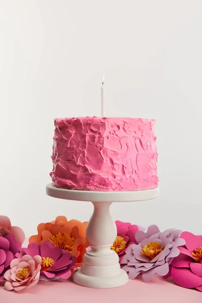 Delicioso pastel de cumpleaños rosa con vela en pie de pastel cerca de flores de papel aisladas en gris - foto de stock