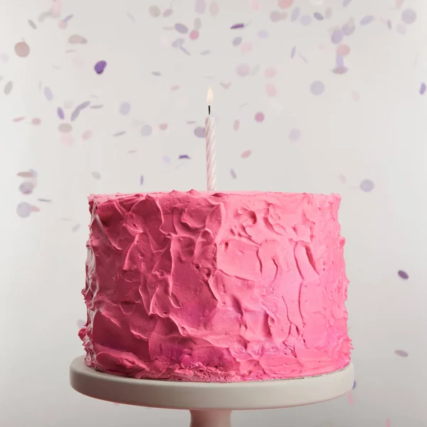 Close up of delicious pink birthday cake with candle on cake stand near confetti on grey — Stock Photo