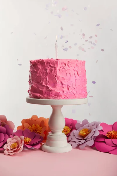Délicieux gâteau d'anniversaire rose avec bougie sur le stand de gâteau près de fleurs en papier et confettis sur gris — Photo de stock