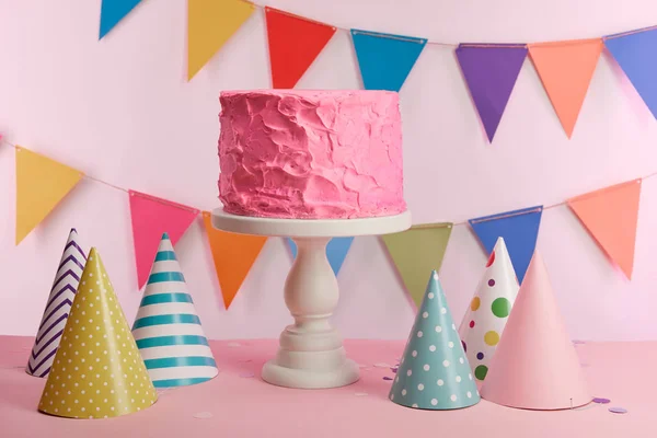 Delicioso pastel de cumpleaños rosa en pie de tarta cerca de gorras de fiesta y decoración - foto de stock