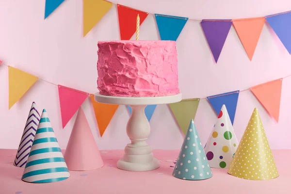 Delicious pink birthday cake with candle on cake stand near party caps and decoration — Stock Photo