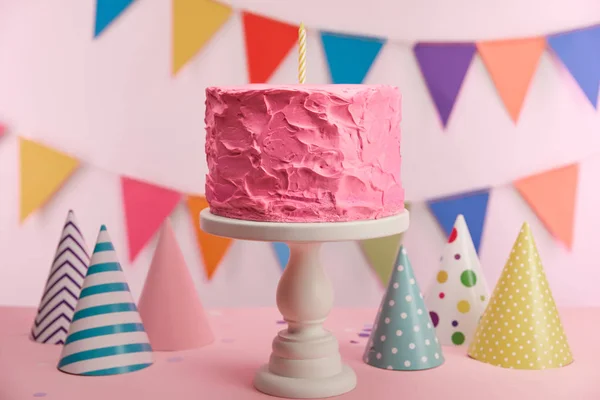 Selective focus of delicious pink birthday cake with candle on cake stand near party caps and decoration — Stock Photo