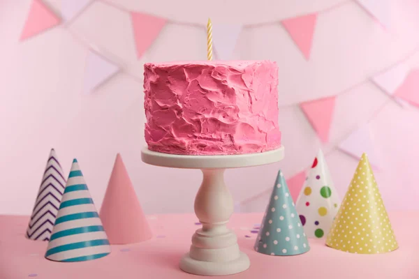 Selective focus of tasty pink birthday cake with candle on cake stand near party caps and decoration — Stock Photo