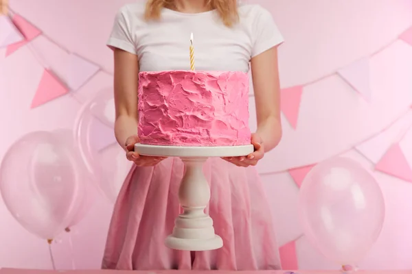 Cropped view of woman holding cake stand with sweet pink birthday cake and burning candle near air balloons and decoration — Stock Photo