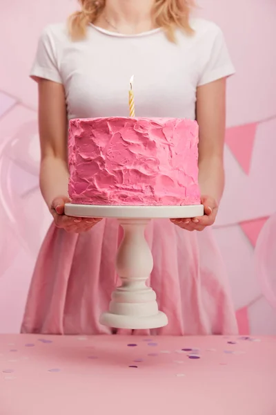 Cropped view of woman holding cake stand with sweet pink birthday cake and burning candle — Stock Photo