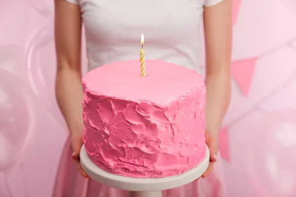 Cropped view of woman holding cake stand with tasty pink birthday cake and burning candle — Stock Photo