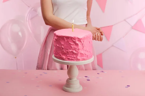 Cropped view of woman standing near sweet and tasty pink birthday cake with burning candle on cake stand — Stock Photo