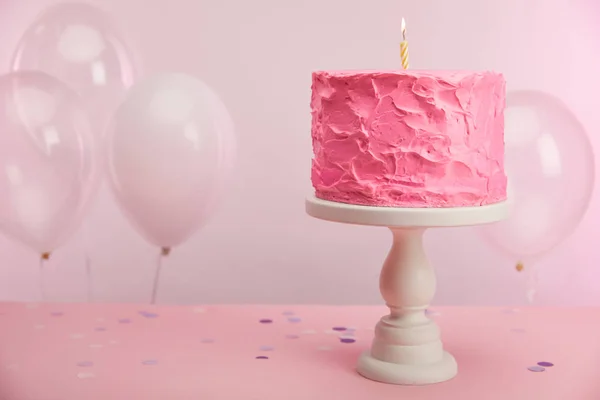 Sweet and tasty birthday cake with burning candle on cake stand near air balloons and decoration on pink — Stock Photo