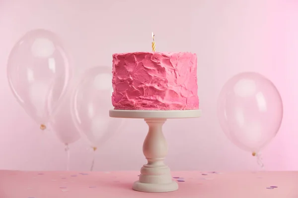 Süße und leckere Geburtstagstorte mit brennender Kerze auf Kuchenständer neben Luftballons auf rosa — Stockfoto