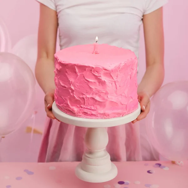 Vista ritagliata della donna che tiene il supporto della torta con gustosa torta di compleanno rosa e candela numero uno ardente vicino ai palloncini d'aria — Foto stock