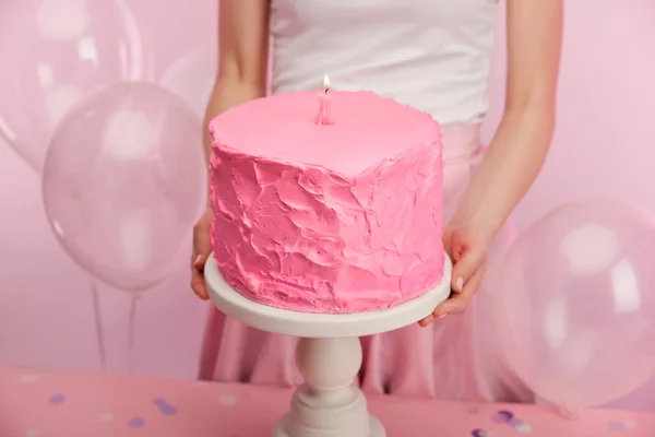 Cropped view of woman holding cake stand with pink birthday cake and burning number one candle near air balloons — Stock Photo