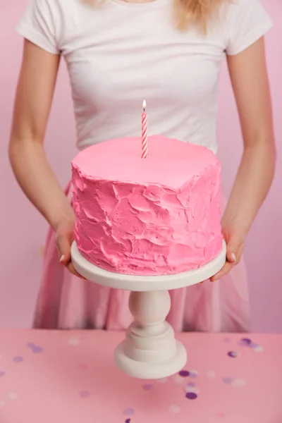 Cropped view of woman holding cake stand with sweet pink birthday cake and burning candle — Stock Photo