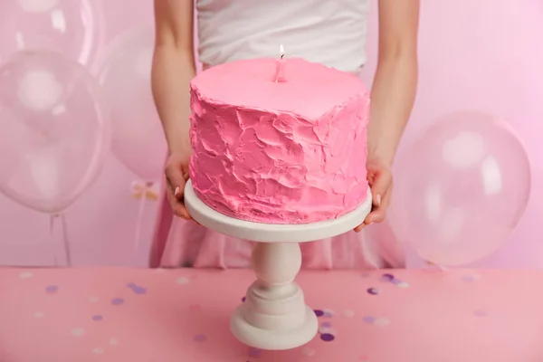 Cropped view of woman holding cake stand with pink sweet birthday cake and burning number one candle near air balloons — Stock Photo