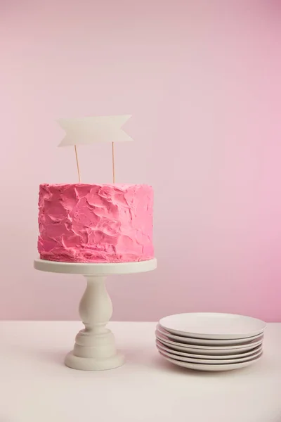 Tasty and sweet birthday cake with blank card on cake stand near saucers on pink — Stock Photo