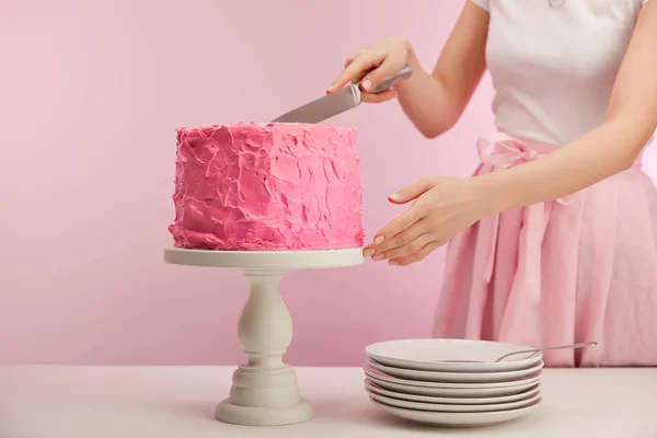 Vue recadrée de la femme tenant un couteau près du gâteau d'anniversaire rose sur le stand de gâteau près des soucoupes sur rose — Photo de stock