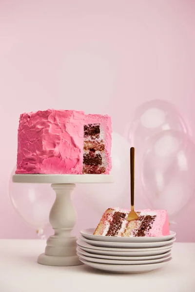 Stück süße Geburtstagstorte mit goldener Gabel in weißer Untertasse neben Luftballons auf rosa — Stockfoto