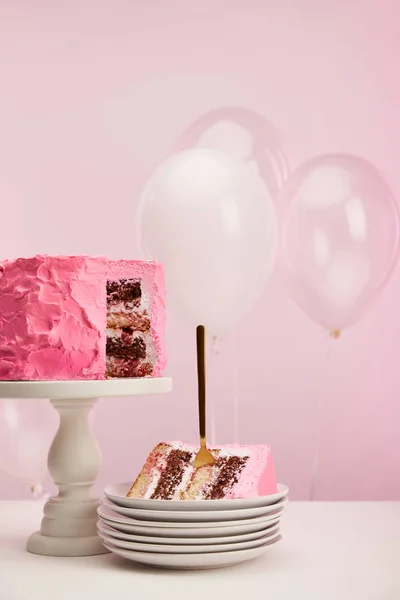 Morceau de gâteau d'anniversaire savoureux avec fourchette dorée dans une soucoupe blanche près de ballons à air sur rose — Photo de stock