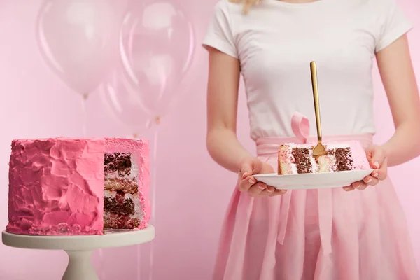 Cropped view of woman holding plate with piece of sweet birthday cake and golden fork on pink — Stock Photo