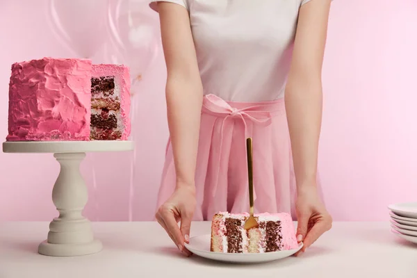 Обрезанный вид женщины, держащей тарелку с кусочком вкусного праздничного торта и золотой вилкой на розовом — стоковое фото