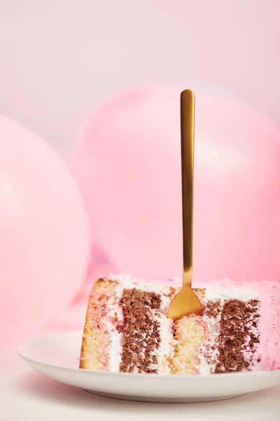 Foco selectivo de dulce pedazo de delicioso pastel de cumpleaños con tenedor de oro en platillo blanco cerca de globos de aire en rosa - foto de stock