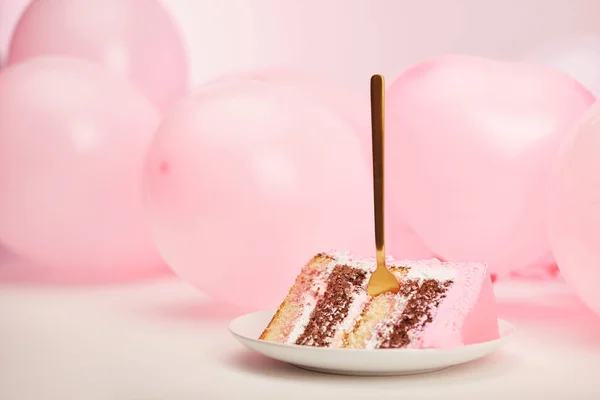 Selective focus of sweet piece of birthday cake with golden fork in white saucer near pink air balloons — Stock Photo