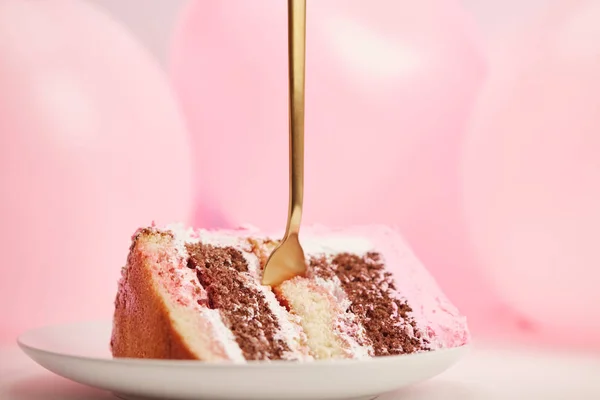 Selektiver Fokus auf leckeres Stück Geburtstagstorte mit goldener Gabel in weißer Untertasse neben rosa Luftballons — Stockfoto