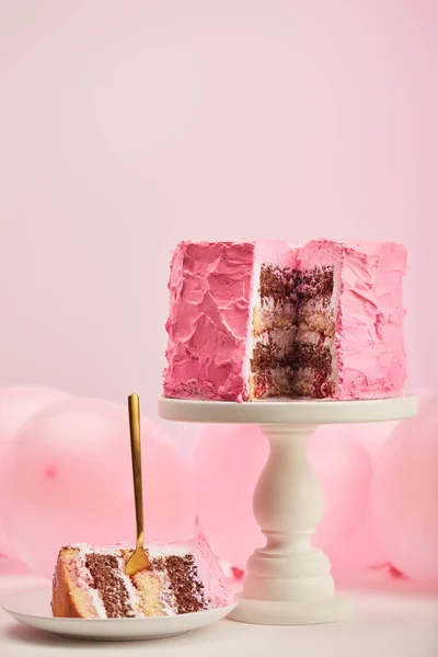 Messa a fuoco selettiva di gustoso pezzo di torta di compleanno dolce con forchetta dorata in piattino bianco vicino a palloncini d'aria e supporto torta su rosa — Foto stock