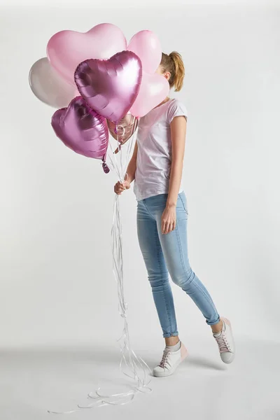 Girl in casual clothes covering face with heart-shaped pink air balloons on white — Stock Photo