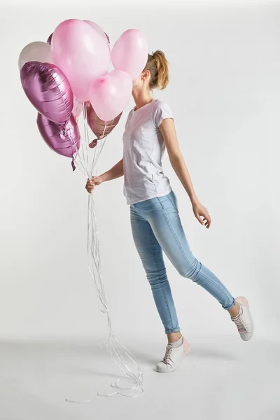 Girl in casual clothes jumping and covering face with pink air balloons on white — Stock Photo