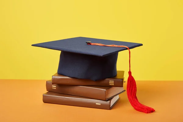 Gorra académica y libros marrones sobre superficie anaranjada aislada sobre amarillo - foto de stock