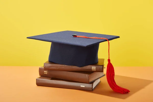 Akademische Mütze und braune Bücher auf orangefarbener Oberfläche isoliert auf gelb — Stockfoto