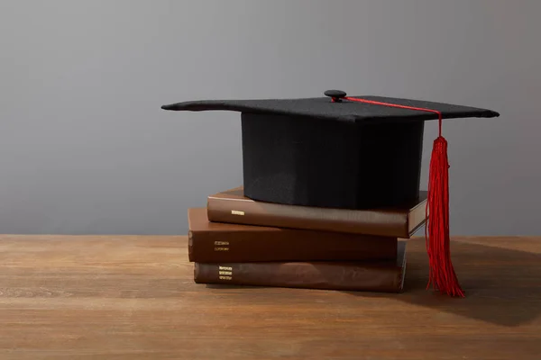 Braune Bücher und akademische Mütze auf hölzerner Oberfläche isoliert auf grau — Stockfoto