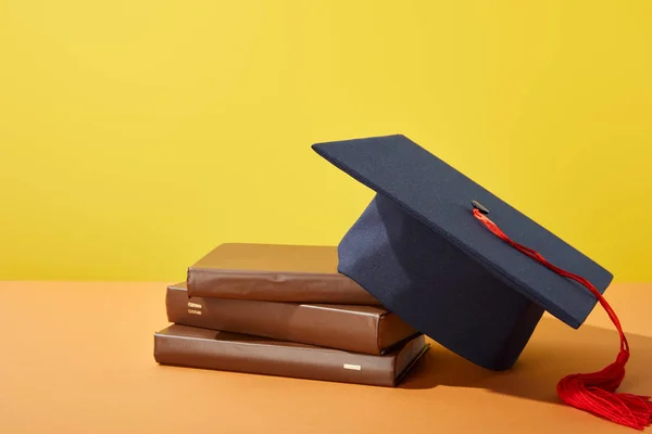 Braune Bücher und akademische Mütze mit roter Quaste auf orangefarbener Oberfläche isoliert auf gelber — Stockfoto