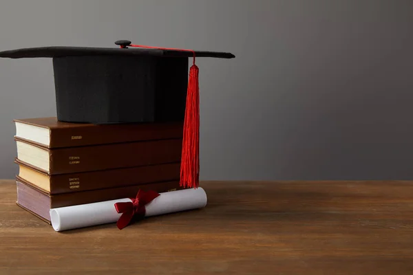 Диплом, академическая шапка и книги по деревянной поверхности на сером — стоковое фото