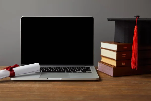 Ноутбук з порожнім екраном, книги, диплом та академічна шапка на дерев'яному столі ізольовані на сірому — стокове фото