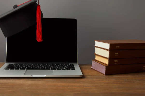 Laptop with blank screen, books and academic cap with red tassel on wooden surface isolated on grey — Stock Photo