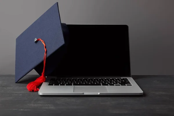 Laptop mit leerem Bildschirm und akademischer Mütze mit roter Quaste auf grau — Stockfoto