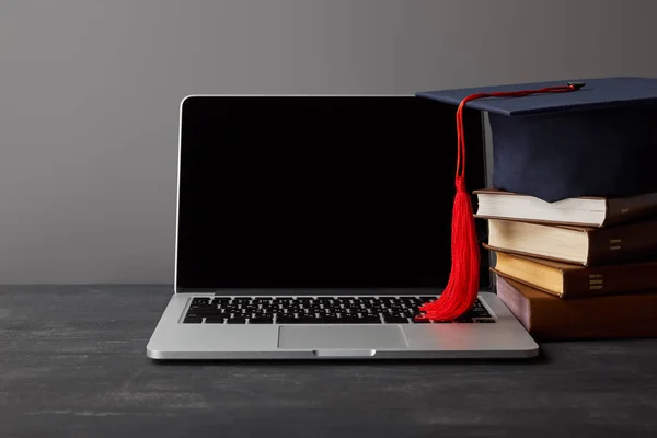 Portátil con pantalla en blanco, libros y gorra académica con borla roja sobre gris - foto de stock