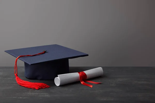 Диплом и академическая шапка с красной кисточкой на темной поверхности на сером — стоковое фото