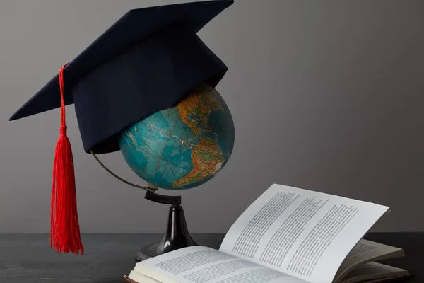 Globe, casquette académique avec pompon rouge et livre ouvert sur la surface de texture sur gris — Photo de stock