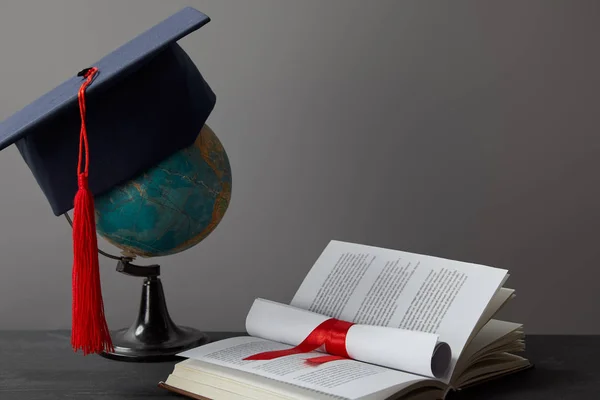 Globo, gorra académica, diploma y libro abierto sobre gris - foto de stock